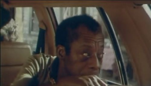 An horizontal image of James Baldwin in the backseat of a car. It is a close crop of Baldwin's face as he is turned around looking out the back window of the car. His right hand appears to hold the head rest, as he holds the left side of his body to look out the car window.