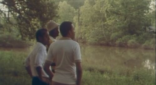 Rectangular image of three black men standing on a grassy, green riverbank. The brownish river splits the image in three, with the other side of the riverbank filled with trees. The men stand in a line, with the middle and far right men fully visible and the head of the far left man visible. That man has a cream white hat on. The two men we can see have their arms on their hips. We cannot see their legs.
