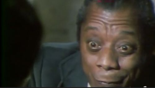 Close crop in color of James Baldwin's face. He looks animated and mid-speech. There is a black shadow that occupies the entire left of the image. It is conceivably the person he's speaking to. The shadow is a semi-circular with an outline consistent with a face.