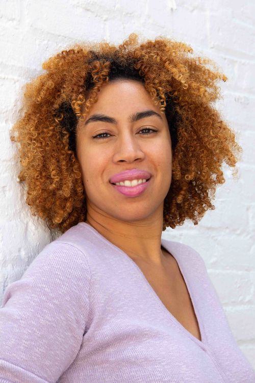 Photo portrait of Jasmin Hernandez, leaning her back against a white brick wall and smiling directly at the camera.