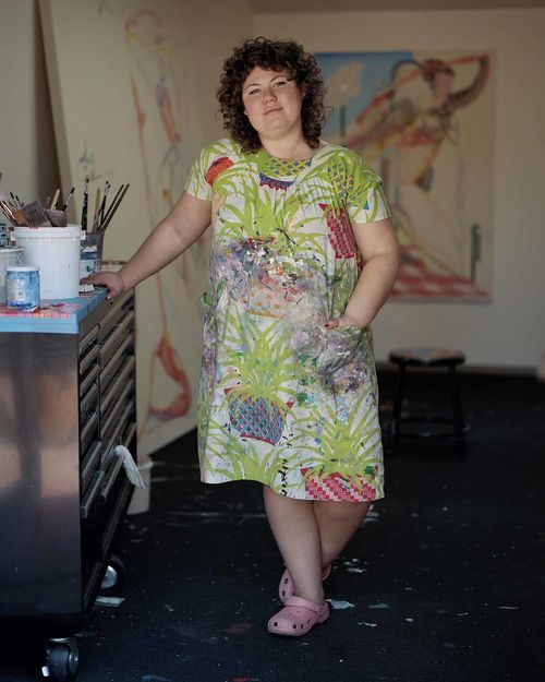 A person with light skin and short curly light brown hair wears a dress with a bright pattern of green and red, and stands in a room with a painiting wall in the background. The person leans an arm on a utility cart on the left of the image, which has tubs filled with paintbrushes on top.