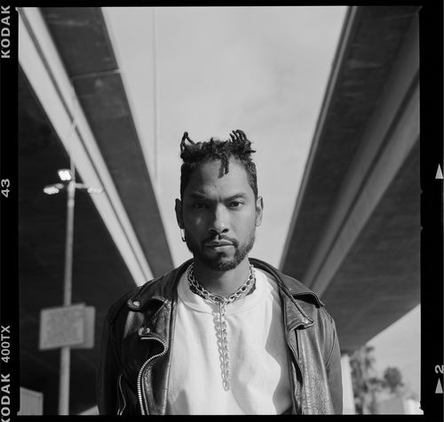 A black and white photograph of a person with a dark beard and short hair, cut in a sculptural way. Seen from the stomach up, the person wears a white t-shirt and leather jacket and stands outdoors, below two concrete freeway flyovers.
