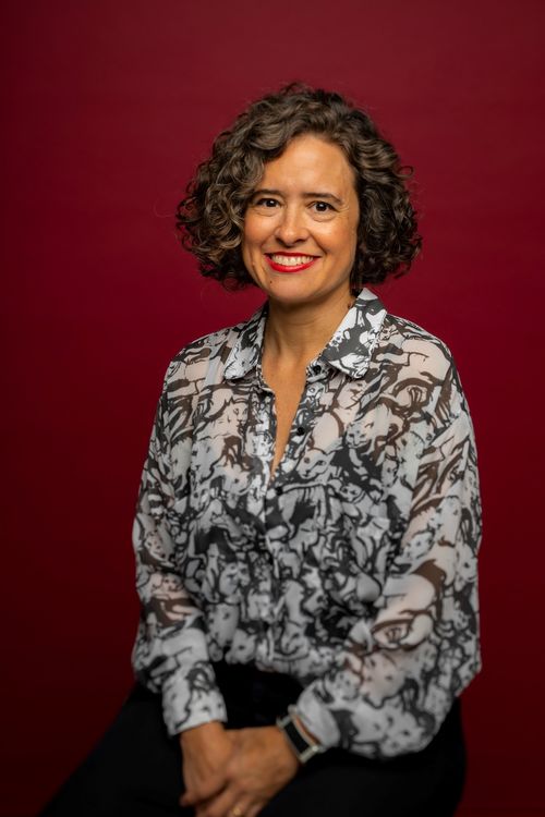 A woman in a flower-pattern blouse sits before a dark red backdrop