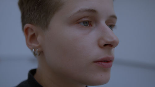 Close up photograph of a person's head in 3/4 view, looking to the viewer's right. The person has light skin, blue eyesand closely shorn hair on the sides, and waers two small silver hoop earrings in the one ear visible.