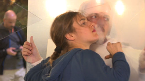 A person in a blue shirt with lng hair in a braid leans a cheek against a transparent surface. Another person wearing a white turban and a white shirt is on the other side of hte surface, leaning a cheek against the surface.