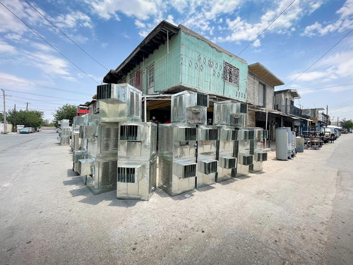 A neat wall of silver air conditioners are stacked three-high on a dusty street with a teal building in the background