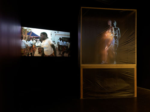 A square structure covered in clear film obscures a figure inside, while a projection plays on the wall to the right