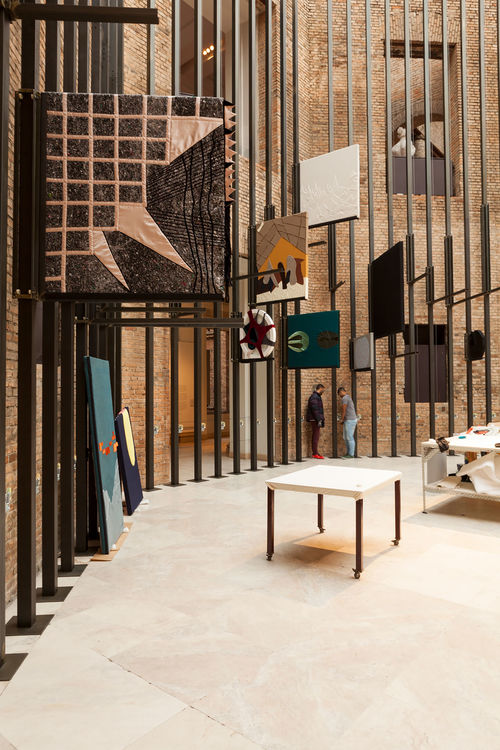 Several textiles hang from a tall wall made of bars, two tables fill the space