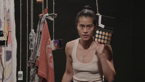 A woman holds up a makeup palette while various objects hang from string around her