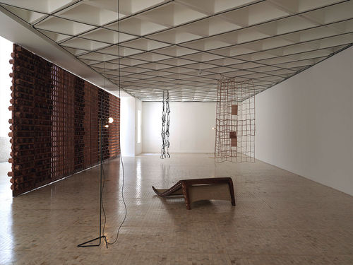 A long gallery room with a chair-like object in the middle of the room and one wall blocked out by a large hanging artwork
