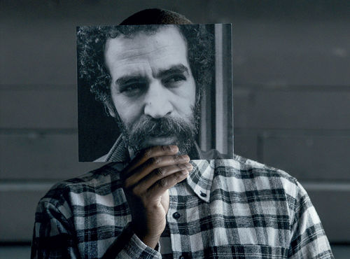 A figure in a plaid shirt holds up a photograph of a face before their face