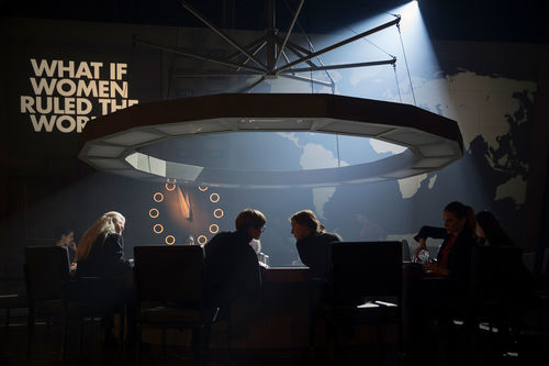 A circle of women around a table with a large circular light fixture above, and the words What if Women Ruled the World on the wall