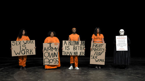Four figures in orange jumpsuits hold up various protest signs, and a grim reaper stands at the end