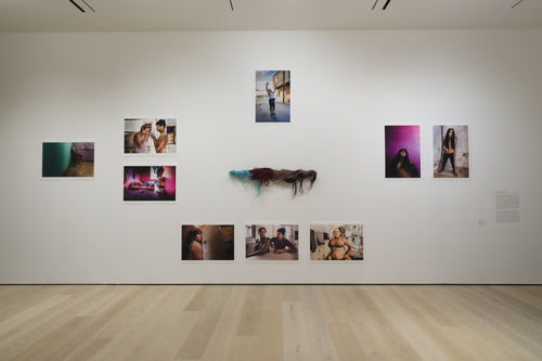Gallery space with photograph of bodies on the wall, with 4 different colored wigs at the center. 