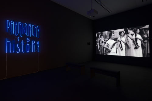 A neon sign that says "Patriarchy is History" is on the wall of a dark room, with a screen featuring a video of several women holding guns beside it.