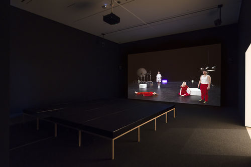 A screen in a dark room shows 4 people wearing red and white lying, sitting, and standing on a dark stage.  