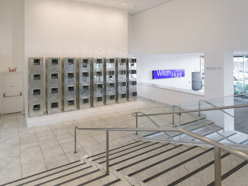 Four rows of silver air conditioners stand stacked on top of each other in a museum lobby. 