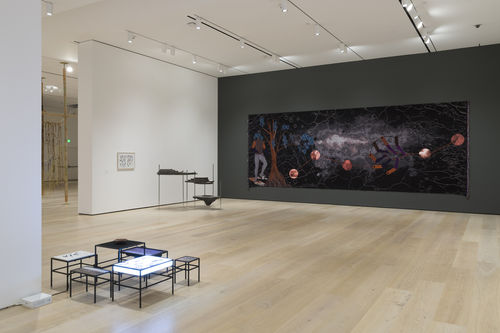 Gallery space with a set of short tiered tables and a textile work that depicts a space scene. 