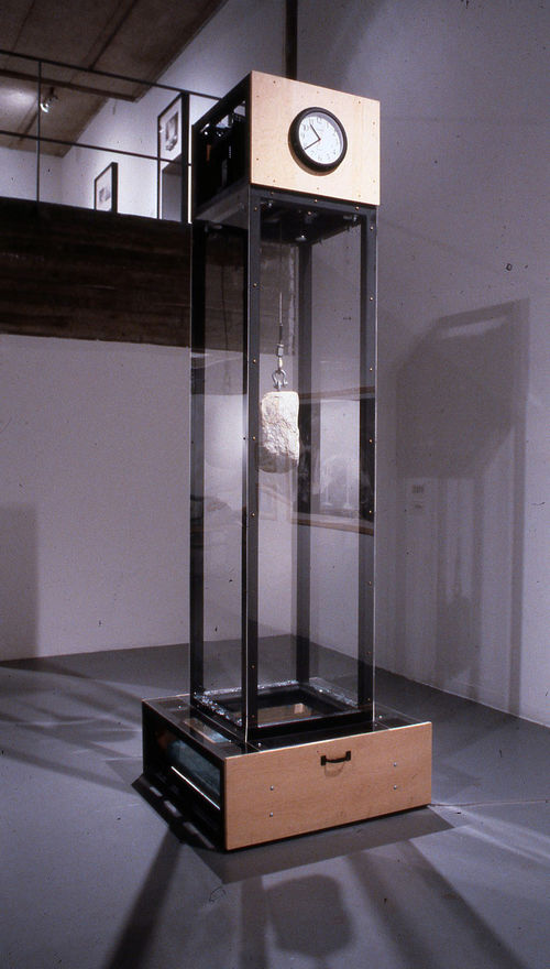 A large rock hung by a wire inside a clear glass box with a clock