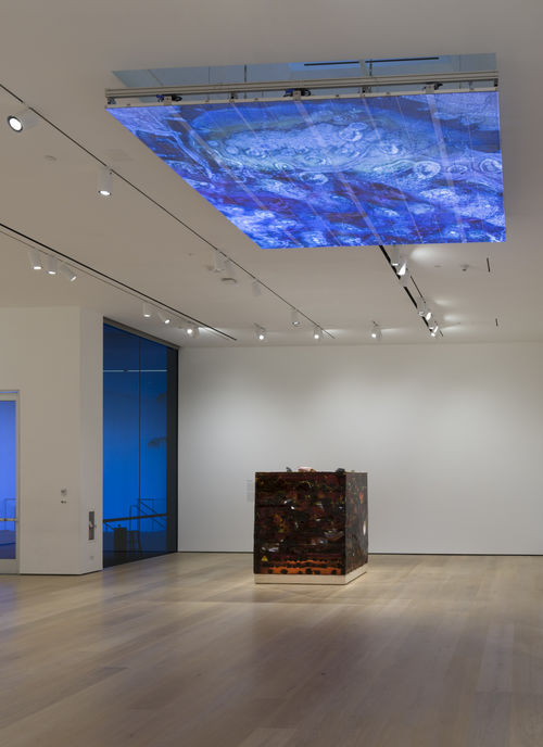 Gallery space with a large amber block in the back, and a large screen on the ceiling showing blue, organic forms.