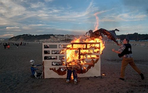 A person swinging a skeleton-like figure into a small structure, which is on fire and contains other skeletons.