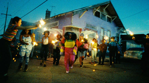 A woman in bright clothes dances and sings, surrounded by a group of people with sparklers