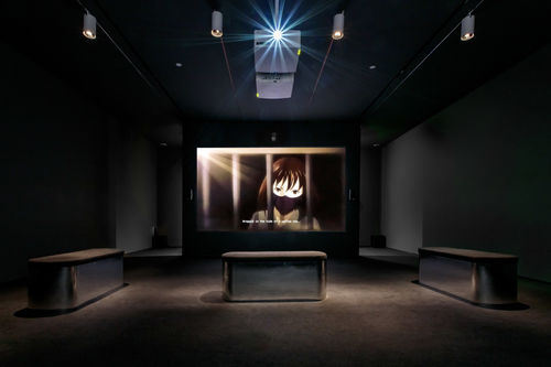 Dark screening room showing video of an anime character in a mask behind bars with the caption "Wrapped in the hide of a yellow cow."