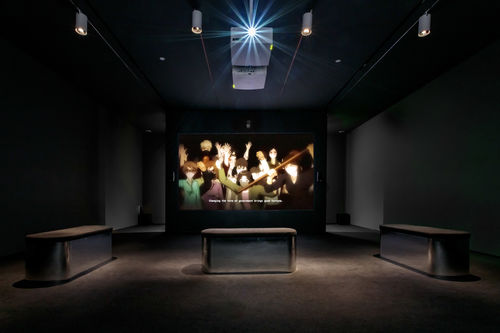 Dark screening room showing video of several anime characters in masks with the caption "Changing the form of government brings good fortune."