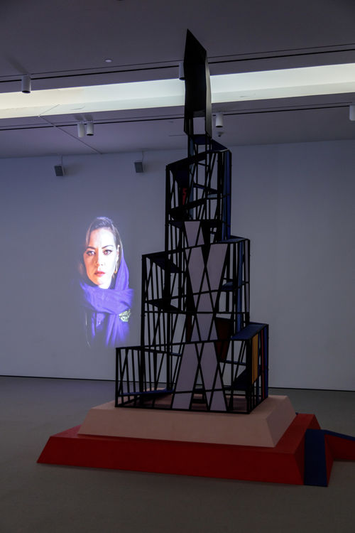 Tall cubist structure with projection of woman in hijab in the background.