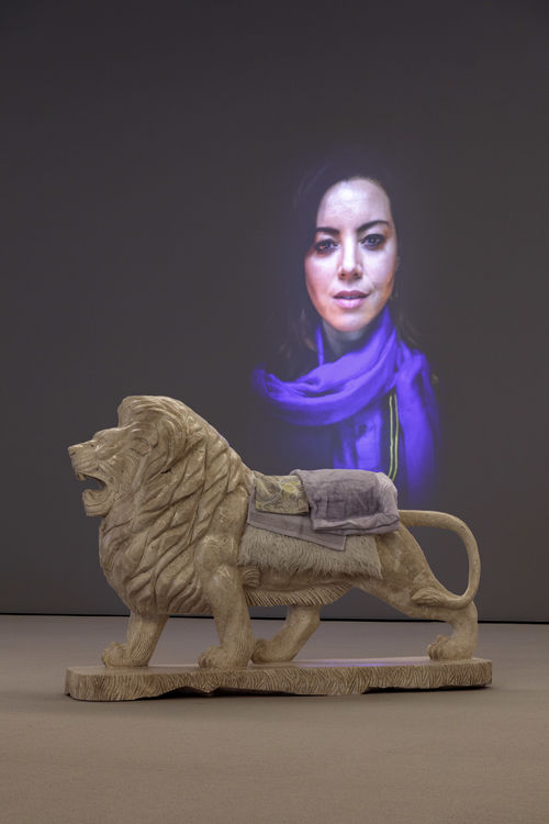 Lion sculpture with textiles laying on top of it in front of projection of woman wearing hijab.