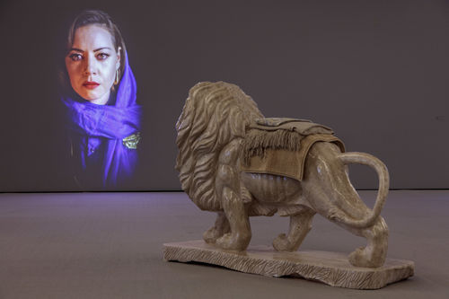 Lion sculpture with textiles laying on top of it in front of projection of woman wearing hijab.