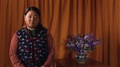 Image of a dark-haired woman next to a vase of purple flowers, in front of a ruffled curtain
