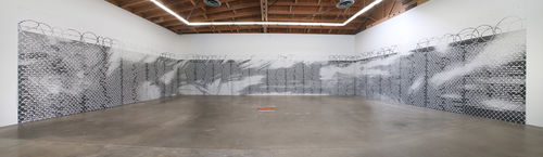 Wide angle view of a large wall piece that resembles a boundary fence covering three walls of the gallery, with barbed wire at the top. The entire fence is covered in names 