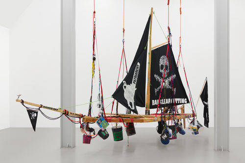 A wooden pirate ship platform hangs from ropes in a gallery. The flags feature feminist and environmental activist symbols and slogans