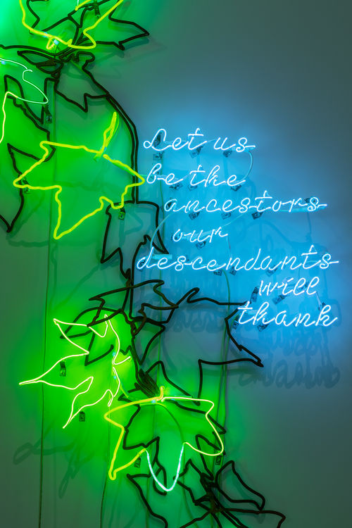 Image of a neon sign of falling leaves and the message, in script, "Let us be the ancestors our descendants will thank"
