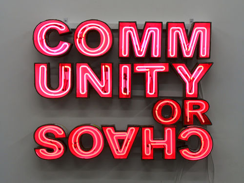 A red neon sign of a block of text: "COMMUNITY OR CHAOS." The word "chaos" upside down
