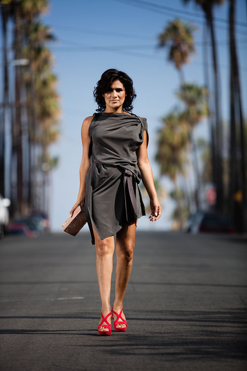 A woman in a grey dress stands in the middle of a palm tree-lined street, holding a brick in her right hand