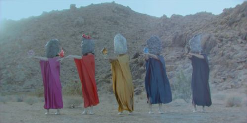 Five people wearing long dresses with their heads covered stand in the hills, holding crystals.