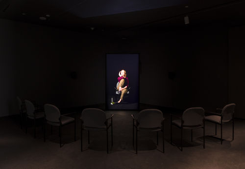 In a darkened room, a semicircle of empty chairs sits in front of a video monitor showing an image of a white woman in a maroon top sitting with finger on temple 