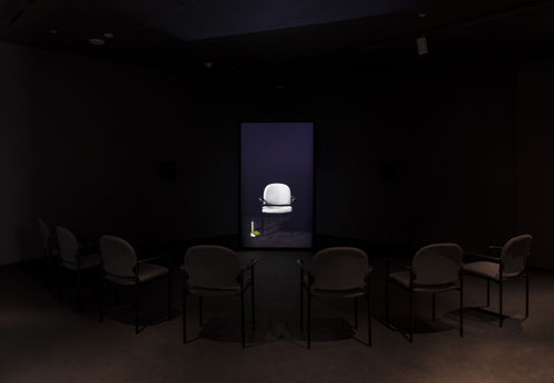 In a darkened room, a semicircle of empty chairs sits in front of a video monitor showing an image of an identical empty chair, with a water bottle and notebook by its side
