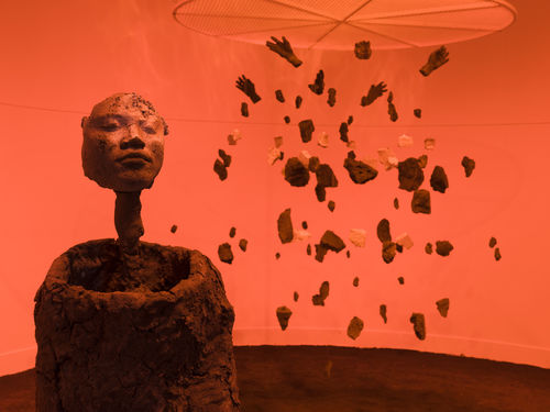 Photo of a sculpture of a form with a human face, and behind it an exploded view of a human figure suspended from the ceiling, in an orange-hued room