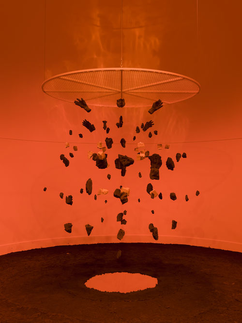 Photo of a sculpture resembling an exploded view of a human figure, suspended from the ceiling in an orange-hued room
