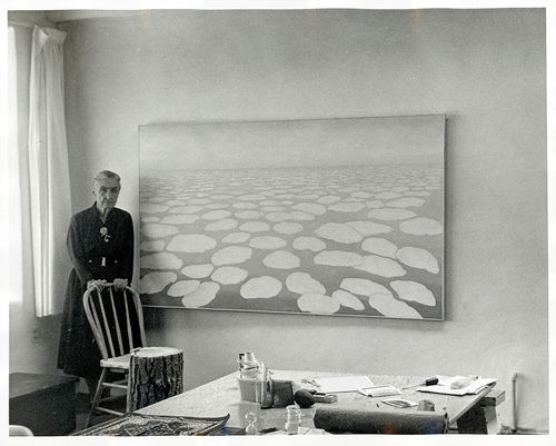 A black and white photo showing the artist Georgia O'Keefe standing in the corner behind a chair, next to a large painting of clouds