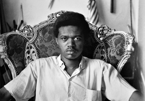 Black and white photograph of a Black man sitting in an ornate chair, looking directly at the camera
