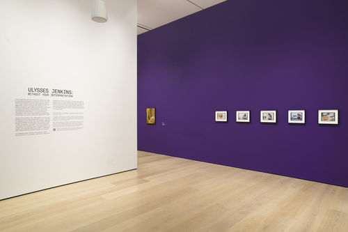Photo of an exhibition showing a series of framed photos on a purple wall, and an introductory text about the exhibition