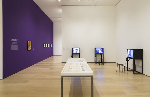 Photo of an exhibition showing framed works on a purple wall, three video monitors, and a table in the middle of the room