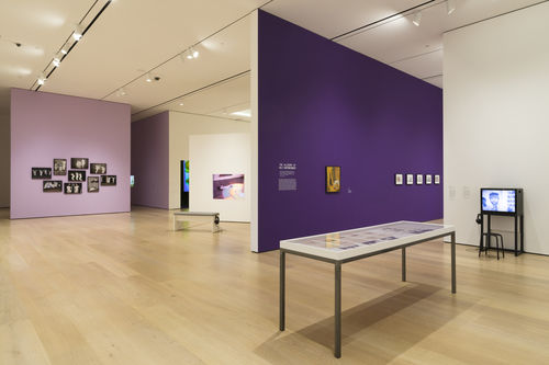 Photo of an exhibition, featuring framed photos, video monitors, and a number of works on a table