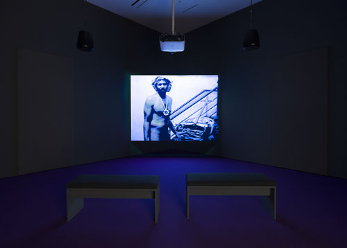 Photo of a video gallery; on the screen the artist stands nude, wearing a large chain around his neck