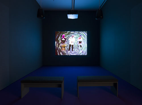 Photo of a video gallery; on the screen three musicians play instruments in front of a psychedelic backdrop