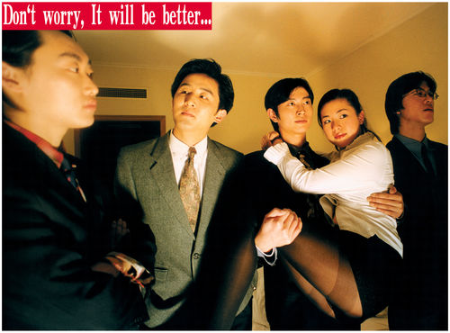 People stand around in an orange lit room. One man holds a woman in his arms